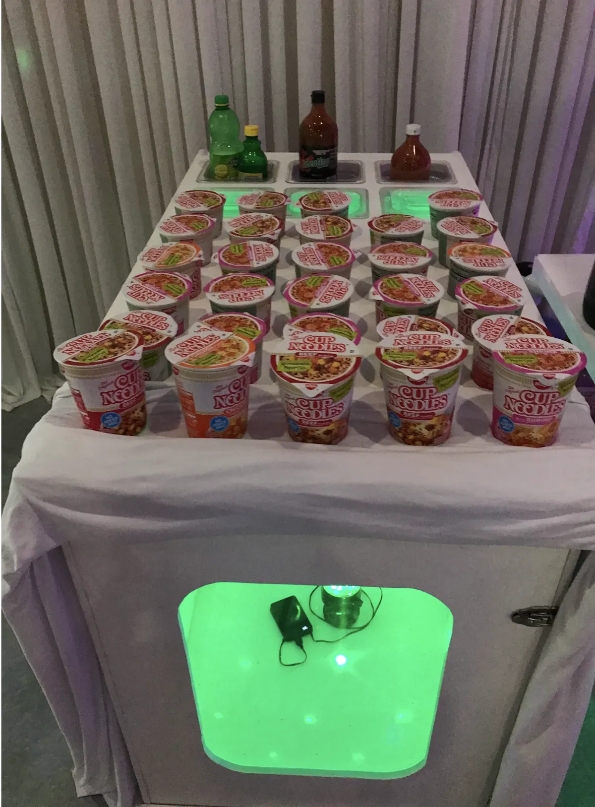 Cup noodle bar with assorted hot sauces and toppings at an indoor event
